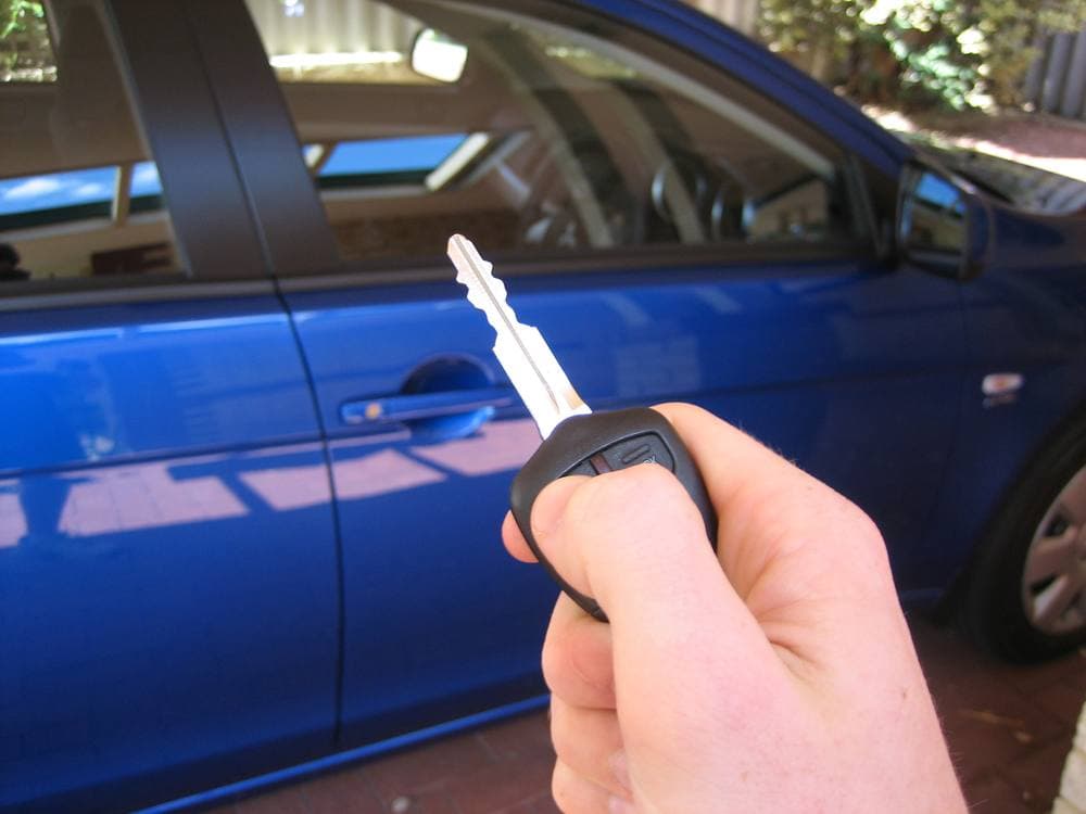 Una mano sostiene la llave con el control de la alarma apuntando hacia la puerta de un carro azul parqueado.